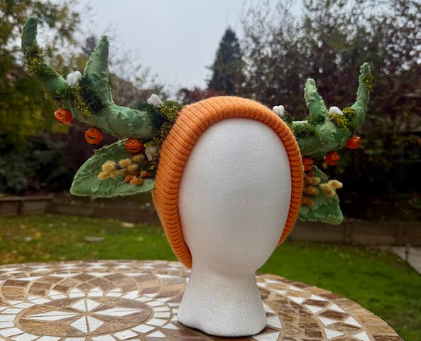 Mossy green antlers on an orange (unlined) beanie. Faux moss, white teeth, and autumnal plants decorate the antlers. 3 jingling orange jack o' lanterns line each antler. Check out the video to hear them chime!