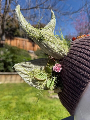 Light & dark brown heather beanie with pale sage green antlers. Gentle light green ferns wrap around each antler with small light pink rose accents. A deep amber jade pendant & beaded chain adorn the front of the beanie for a natural flare.